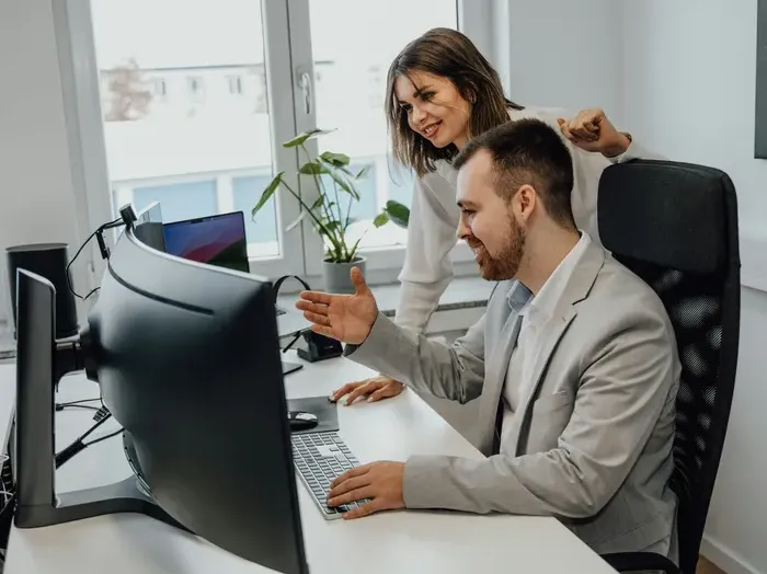 Two coworkers reviewing data on a wide monitor in a bright office, discussing steps and pointing at the screen